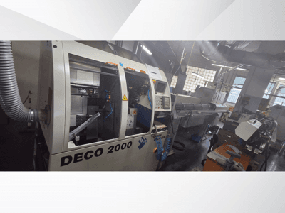 Máquina CNC DECO 2000 en un taller, vista frontal, mostrando los controles y una configuración de pieza larga a la derecha.