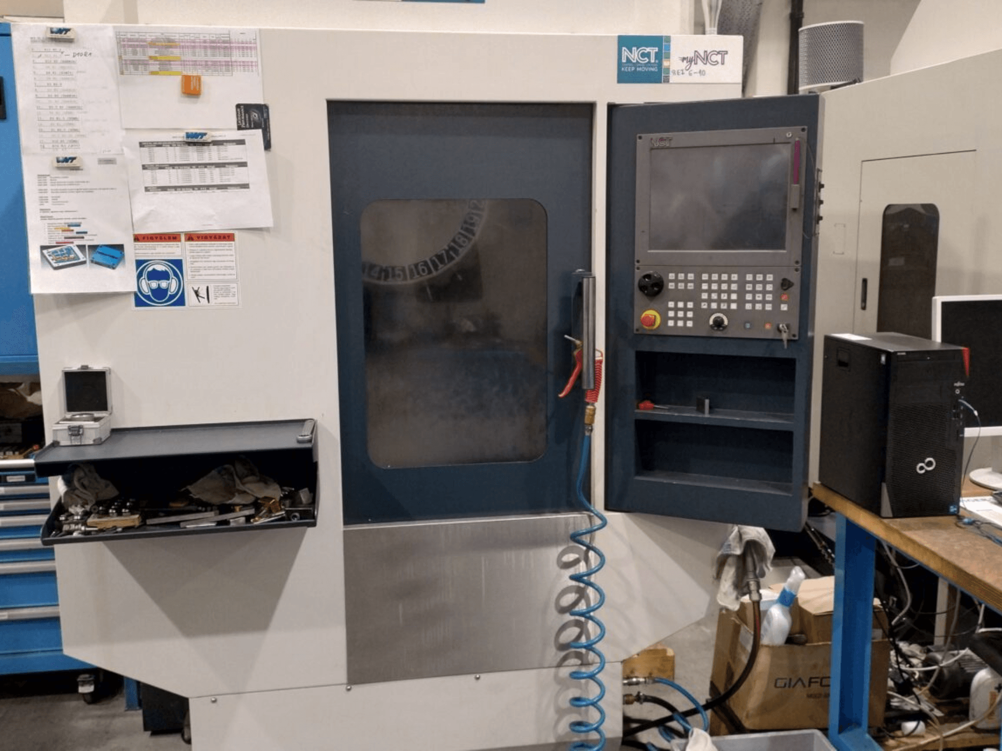 NCT CNC machine, front view, features control panel with numeric keypad, tools in storage drawer, and manual on the wall.