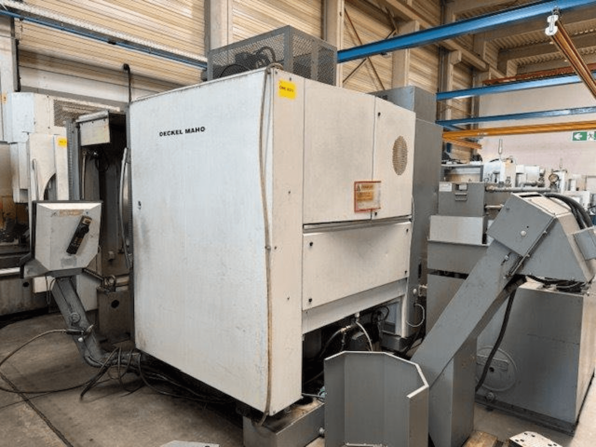 Centro de mecanizado vertical DECKEL MAHO DMC 100 V, vista lateral con panel de control y varios accesorios visibles.