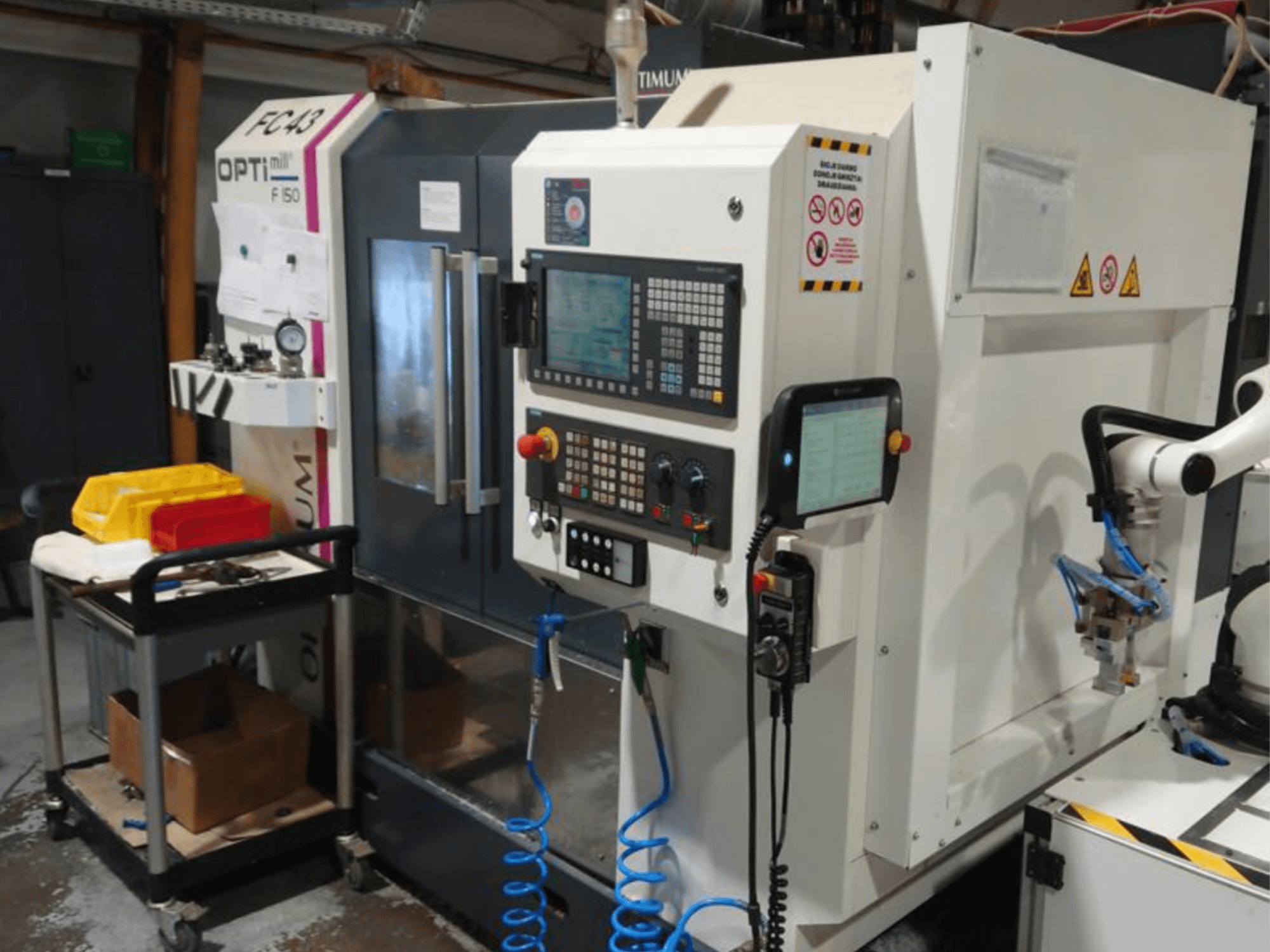 Panel de control de la fresadora OPTIMUM F50 con mangueras, herramientas y un brazo robótico visible en un taller.