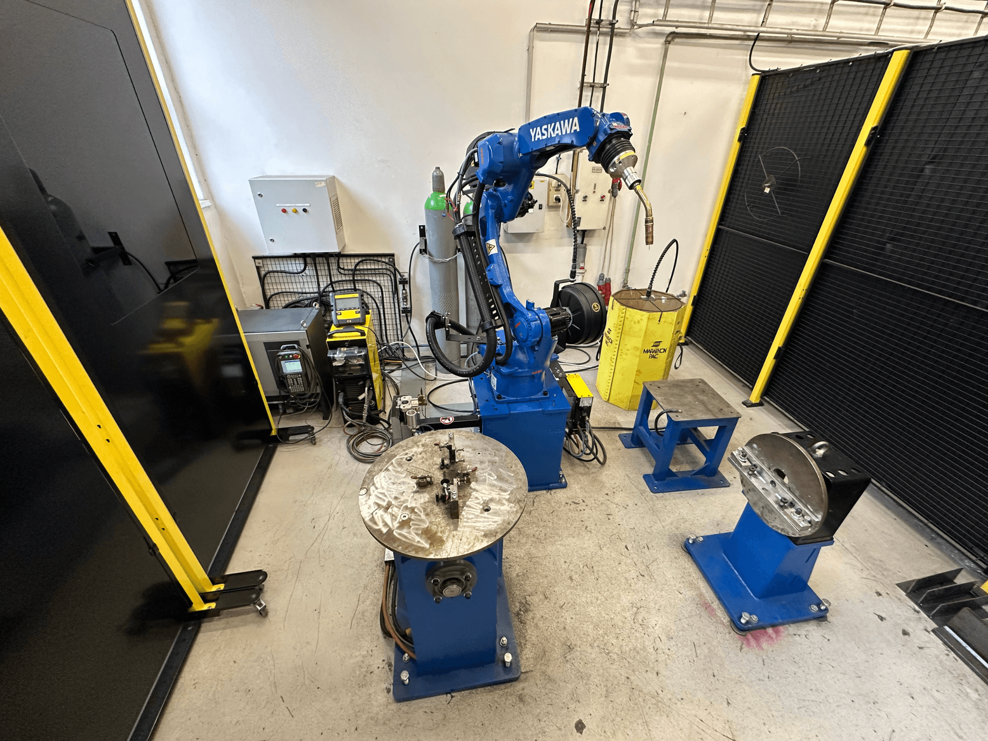 Brazo robótico Yaskawa en azul con dos mesas de trabajo, equipo de soldadura y barreras de seguridad; vista frontal de espacio de trabajo industrial.