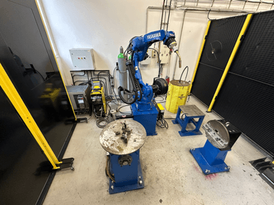 Brazo robótico Yaskawa en azul con dos mesas de trabajo, equipo de soldadura y barreras de seguridad; vista frontal de espacio de trabajo industrial.