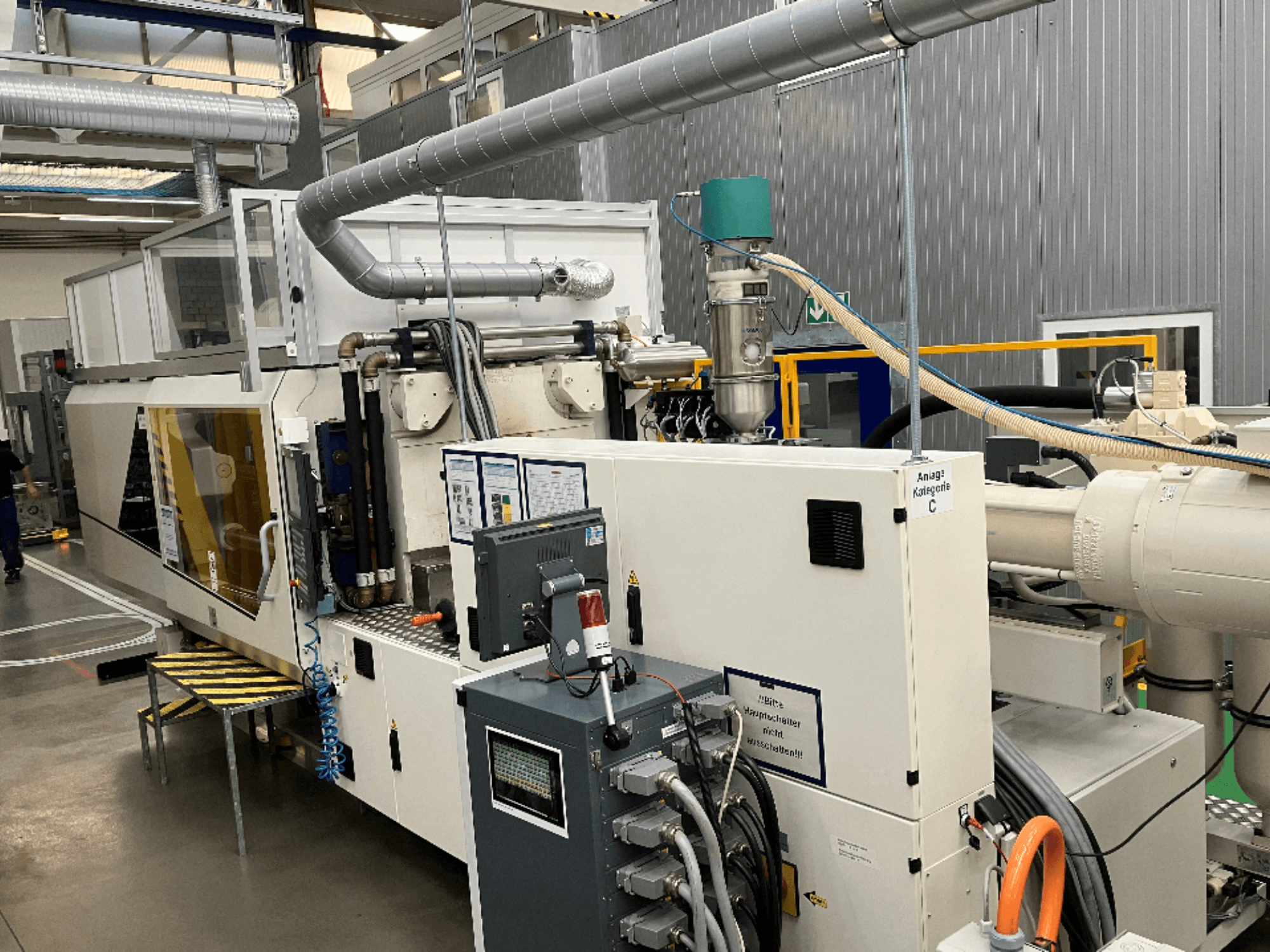 Máquina de moldeo por inyección, vista frontal, con etiqueta de marca, panel de control y mangueras visibles en un entorno de fábrica.