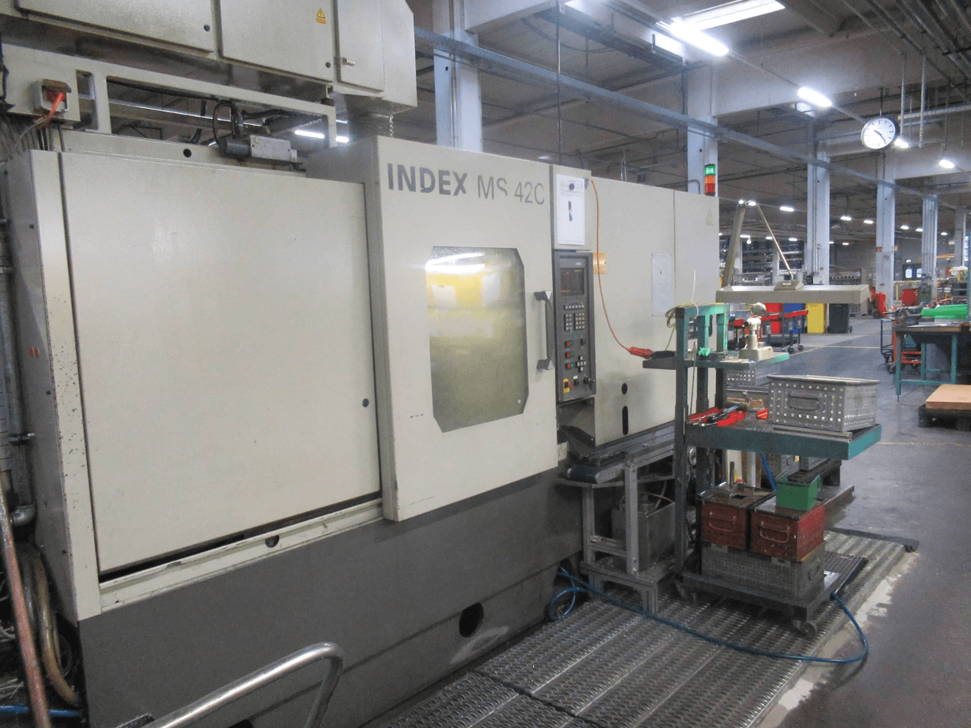 Máquina CNC INDEX MS 42C en un taller, vista lateral que muestra el panel de control y el almacenamiento de herramientas en un carro con ruedas.