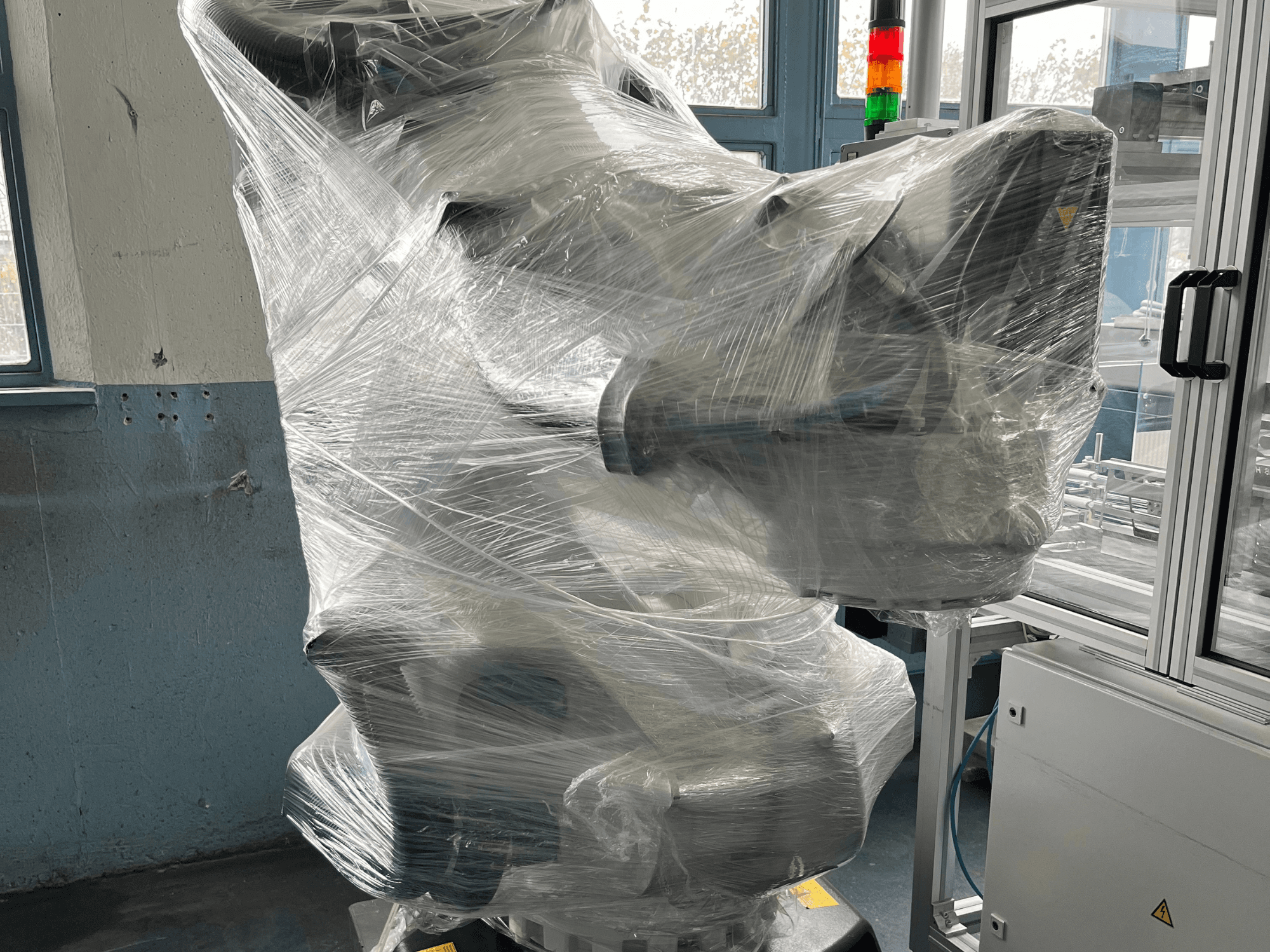 Brazo robótico industrial envuelto en plástico, vista frontal. Incluye unidad de control y luz indicadora. Marca no visible.