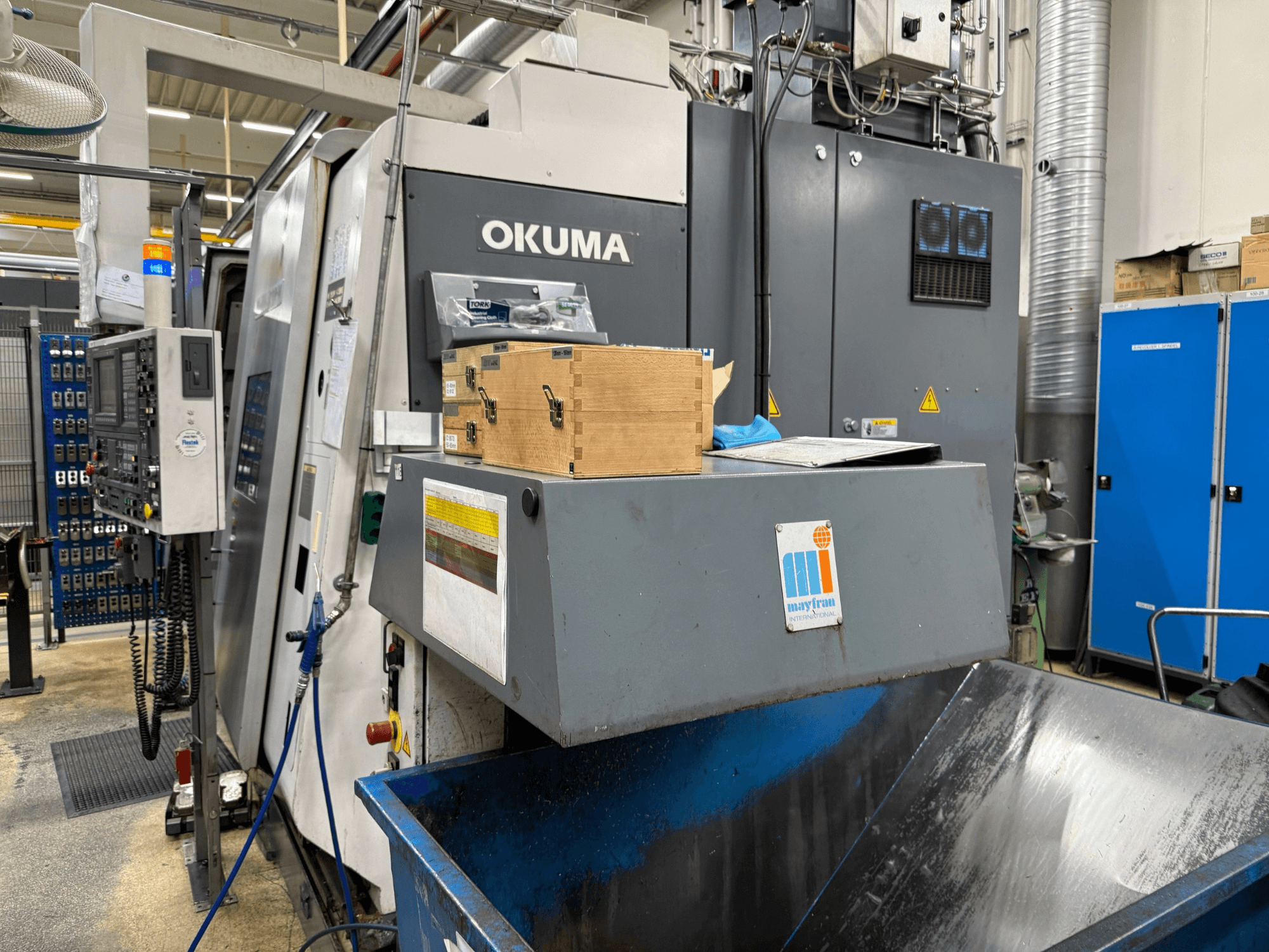 Máquina CNC Okuma en fábrica, vista frontal; cuenta con panel de control, cajas de madera y papelera azul con el logotipo de Mayfran.