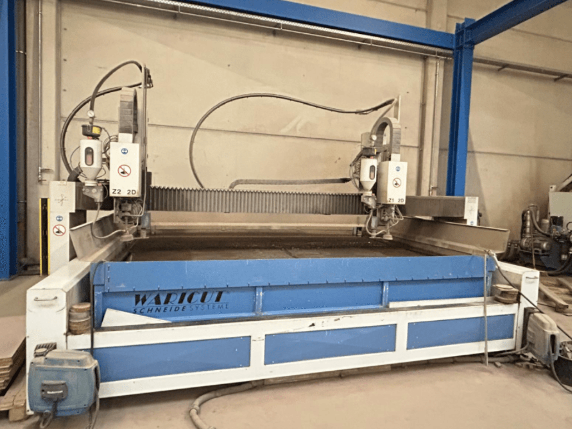 Water jet cutting machine by Waricut, front view; features dual cutting heads and blue work surface in a workshop setting.