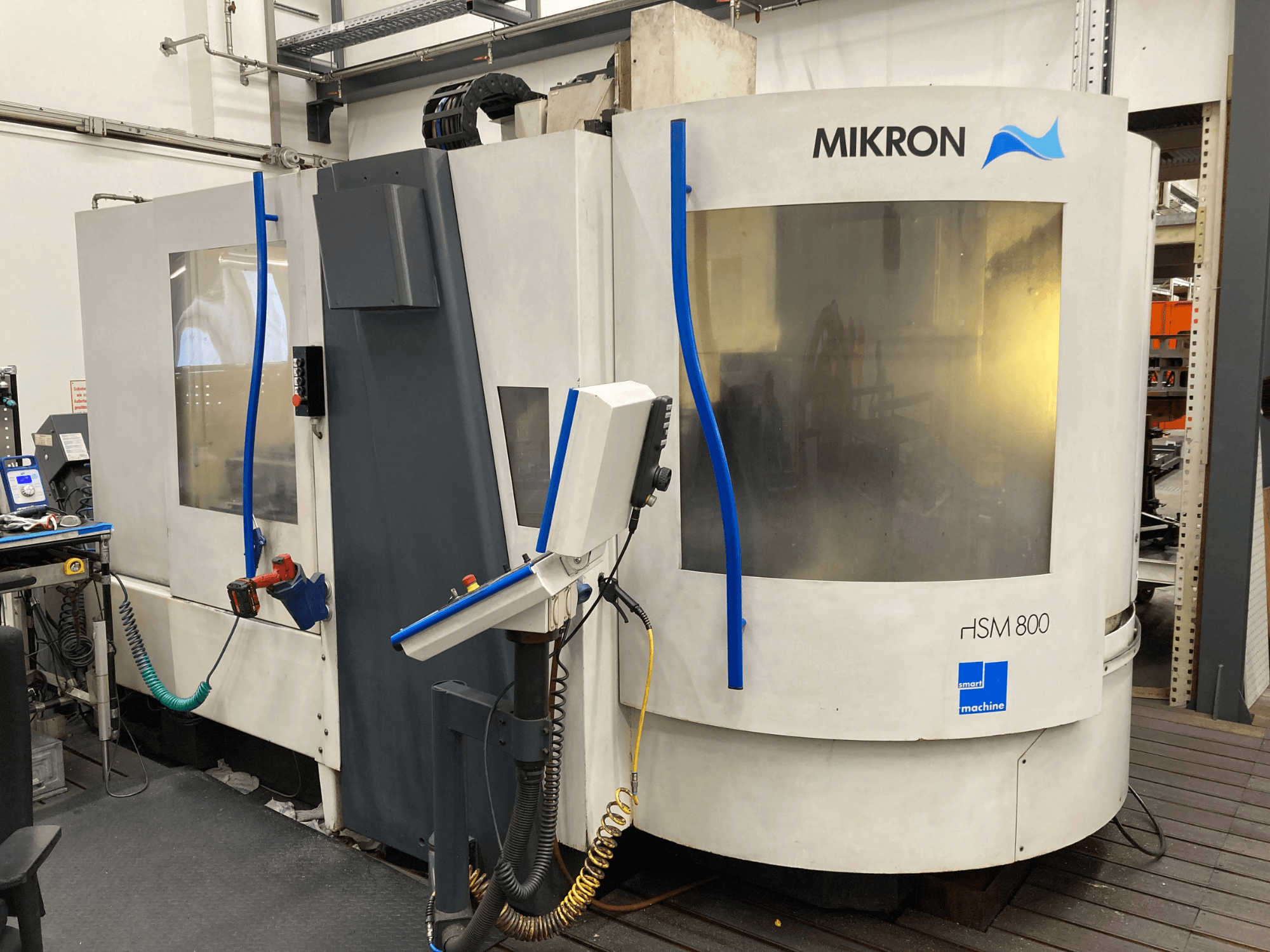 Mikron HSM 800, vista lateral de un centro de mecanizado CNC con panel de control y portaherramientas, mangueras y cables visibles.