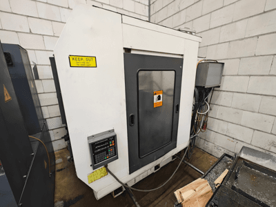 Máquina CNC industrial, vista lateral; presenta panel de control, señales de advertencia y piezas de madera sobre una superficie de trabajo.